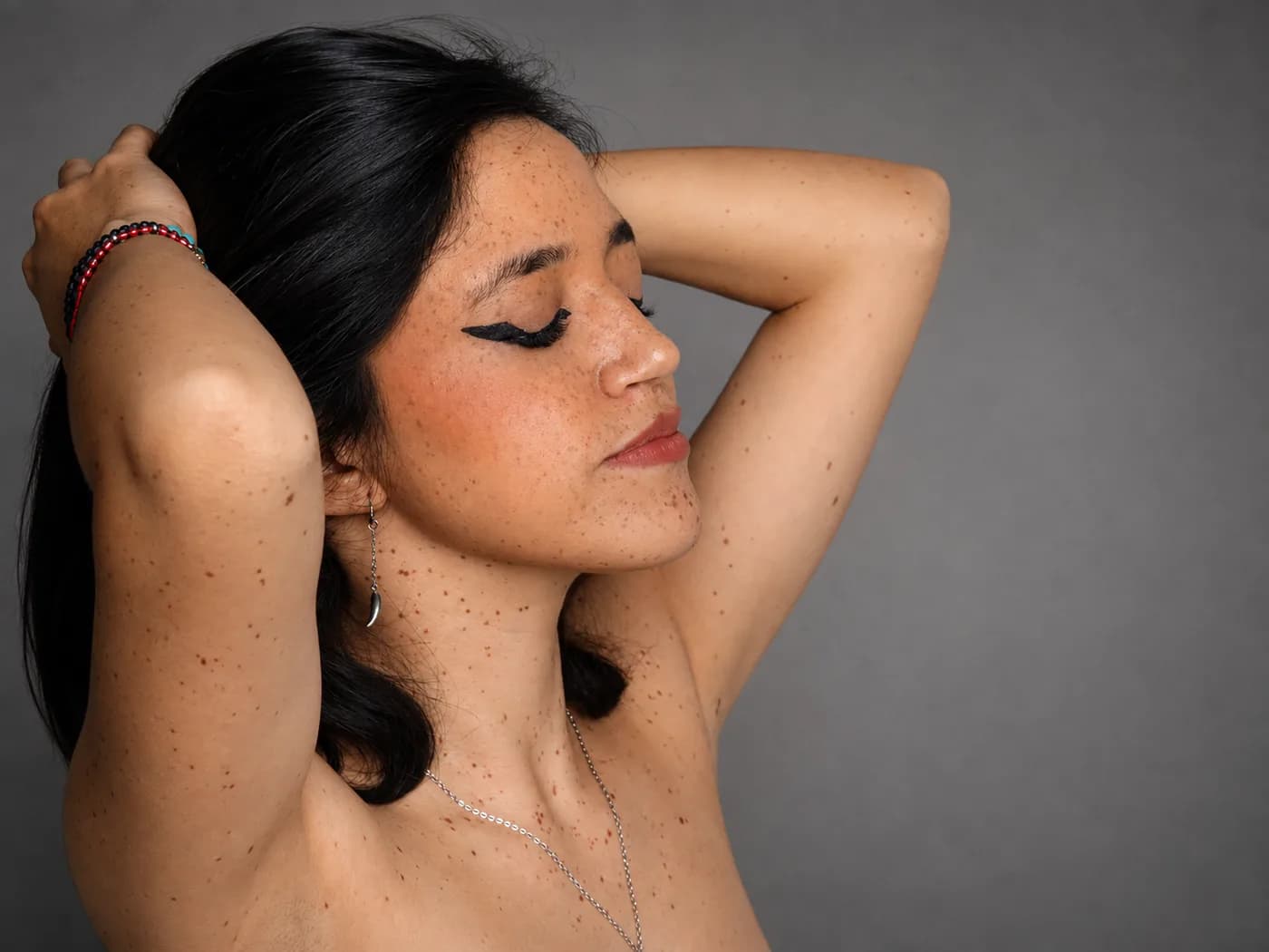 Mujer con delineado gráfico y pecas, los brazos recogidos sobre la cabeza y los ojos cerrados